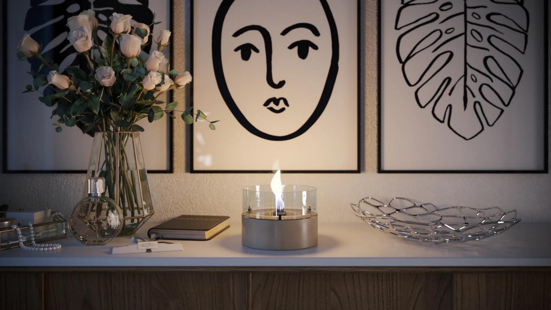 A cylindrical burner on a pale sideboard beneath framed drawings, with roses to the left and a reflective open metal bowl to the right.