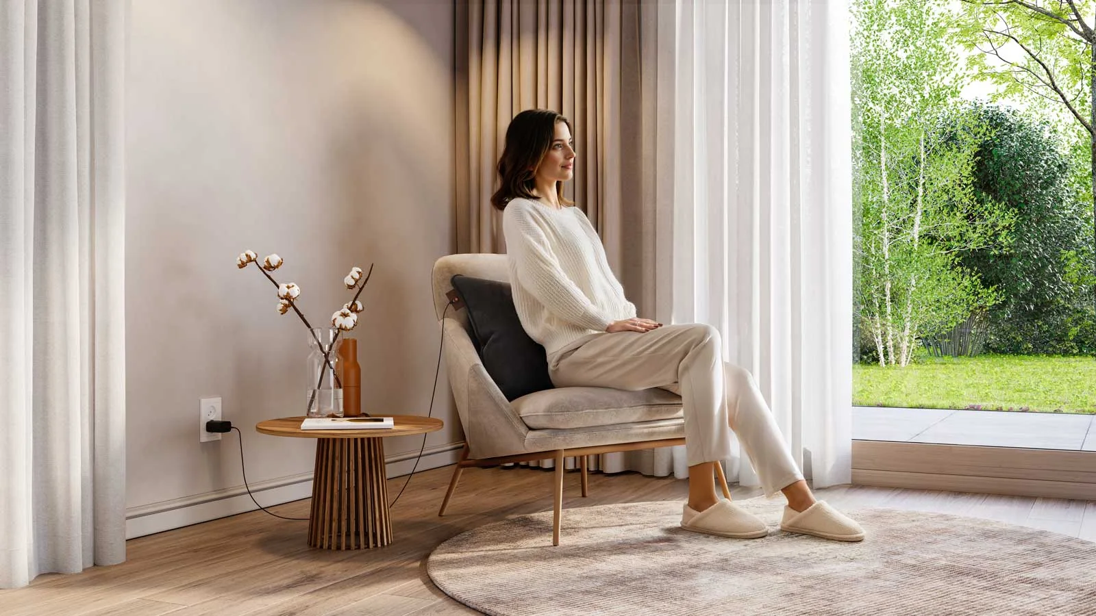 A person sits in a pale armchair beside a garden window, with a dark pillow partly visible behind the back, a knitted sweater and tall curtains nearby.