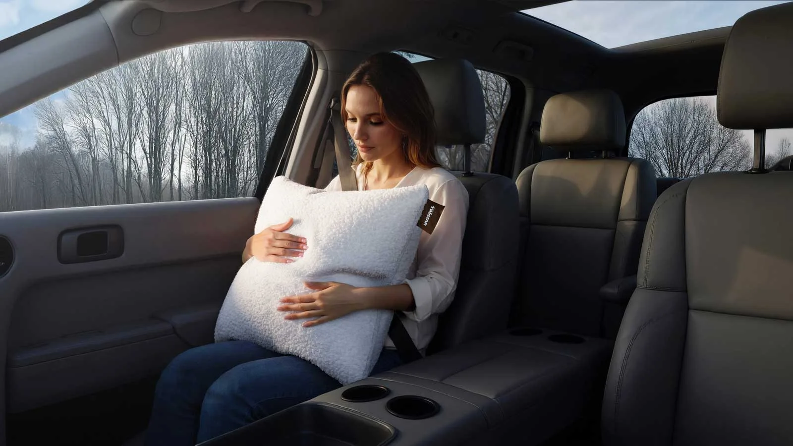 A person in the rear seat of a car holds a cream pillow with a nubby surface and a broad indentation across its middle.